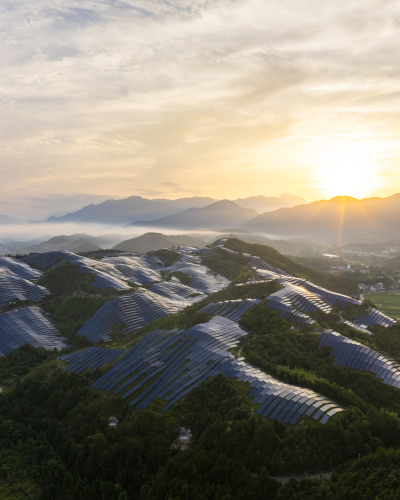 Solar Farms - Carousel Aerial view of a large solar power installation covering rolling hills at sunrise, with rows of solar panels set among green vegetation and mountains in the distance.